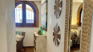 Um corredor com piso de azulejos, uma cadeira de vime, uma cômoda pequena branca e um espelho com moldura trançada.
