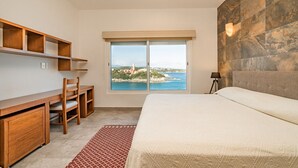 A bedroom with a large bed, a desk, and a chair. There is a window with a view of the sea and buildings. - Oceanfront Oasis Close to Beach w/ Two Pools (Huatulco)