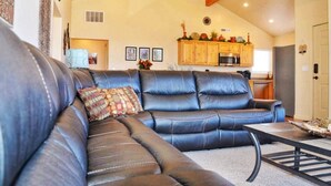 A living room with a sectional sofa, a kitchen in the background, and a high ceiling with exposed beams.