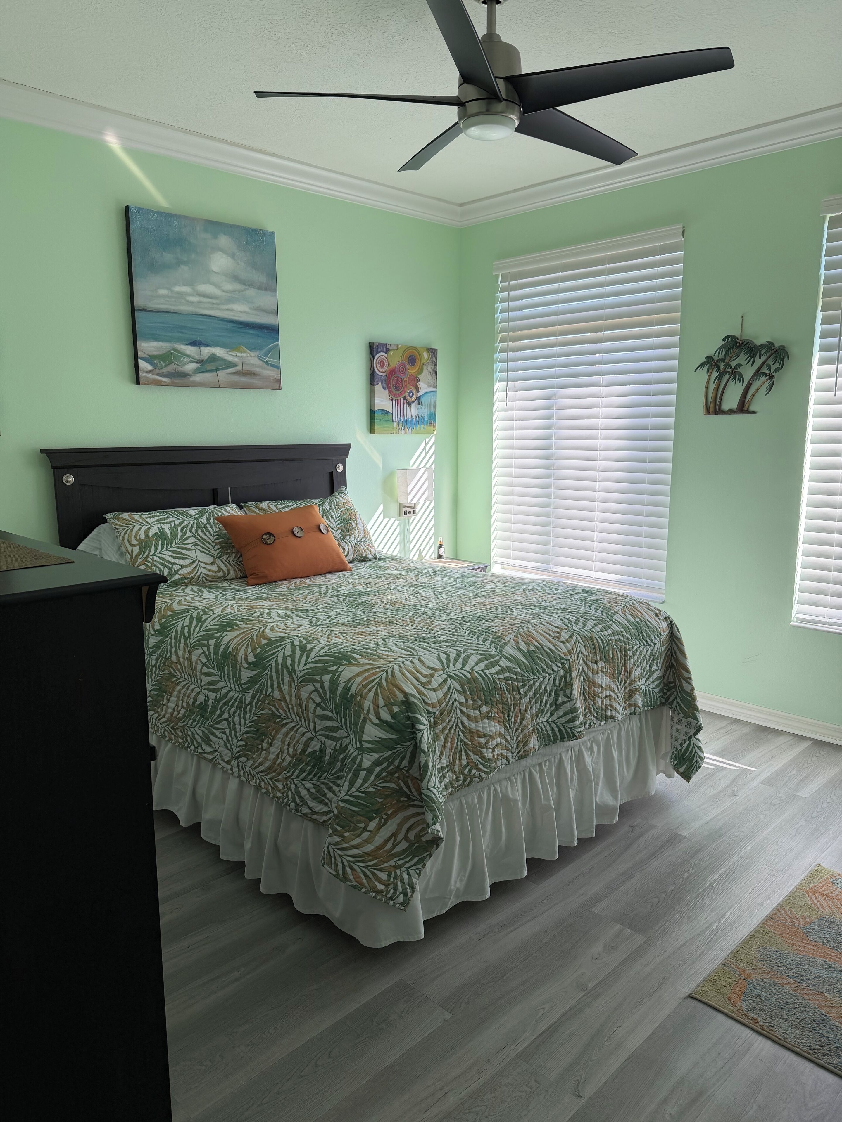Een slaapkamer met een bed, een plafondventilator, een schilderij en een raam met jaloezieën.