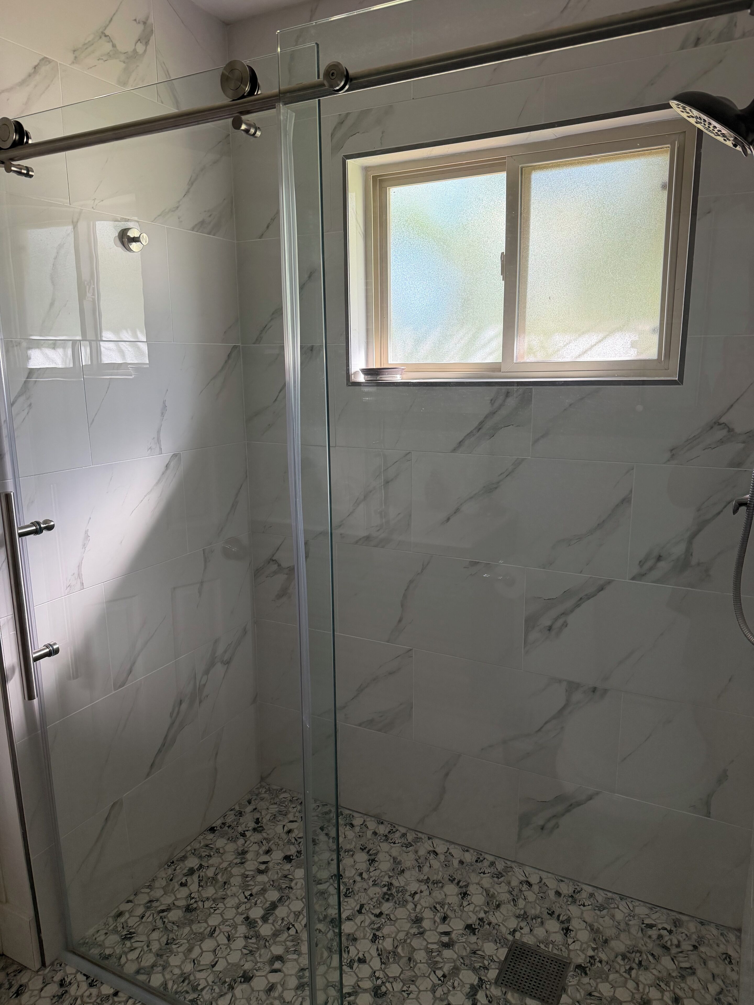 A glass-enclosed shower with a window, marble tiles, and a hexagonal patterned floor.