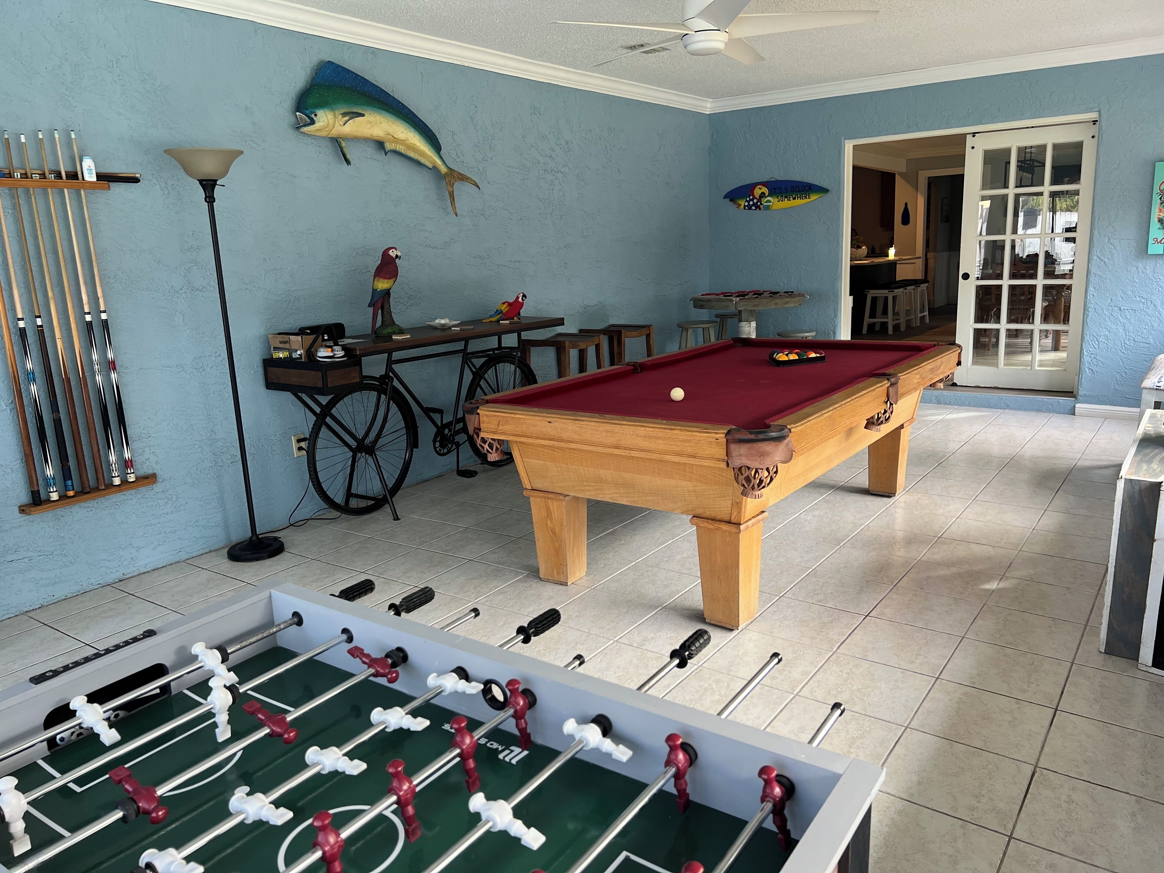 Una mesa de billar, una mesa de fútbol de mesa y equipo de billar en una habitación con paredes azules y un ventilador de techo.