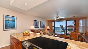 A modern kitchen with a built-in stove, wooden cabinets, and a dining area with a balcony view. - SUMMER SALE! Oceanfront Condo w AC, Ocean Views & Heated Pool - Kaleialoha 309 (Lahaina)