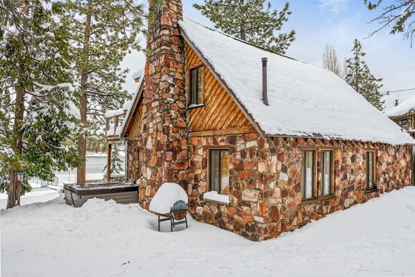 Une cabane en pierre, dont le toit et les fenêtres sont recouverts de neige, entourée d’arbres.