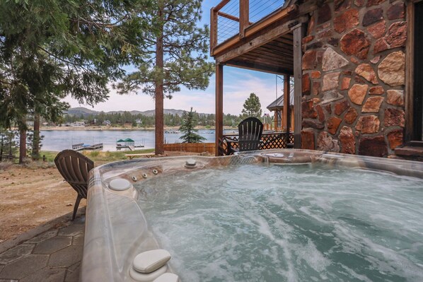 A hot tub with a view of a lake and mountains, a wooden deck, and a stone building.