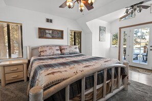 Un dormitorio con una cama grande, cabecera de madera, una mesita de noche, un ventilador de techo y vistas al exterior a través de un ventanal corredero.
