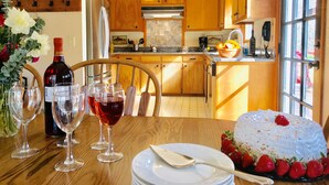 Une cuisine avec des meubles en bois, un réfrigérateur, un évier et une table à manger, mise avec des verres à vin, des assiettes et un gâteau.