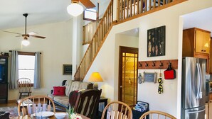 Un coin repas avec une table en bois dressée pour quatre personnes, des chaises, un ventilateur de plafond et un escalier menant à un niveau supérieur.