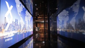 A modern hotel corridor with digital displays on the walls, a glass door entrance, and a reflection of the cityscape.