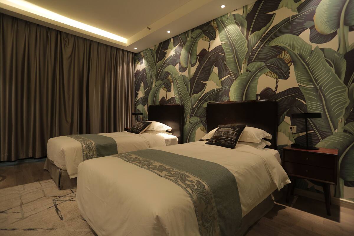 Habitació d'hotel amb dos llits, una finestra gran amb cortines, una paret amb dibuix de fulles tropicals i una meseta de nit amb una làmpada.