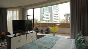 A modern hotel room with a large bed, a TV on a wooden cabinet, a balcony with a view of the city, and a vase with yellow flowers.
