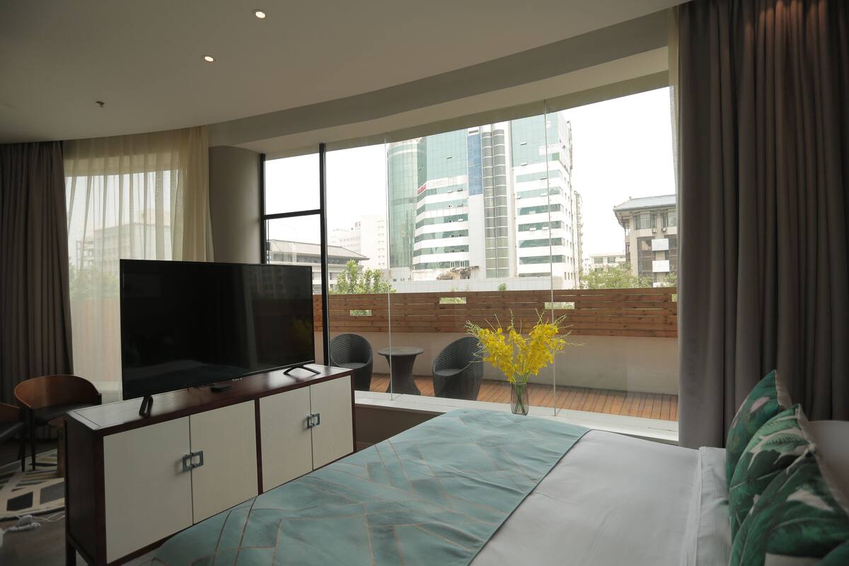 Habitació d'hotel moderna amb un llit gran, una televisió sobre un moble de fusta, un balcó amb vista a la ciutat i un ram de flors grogues.
