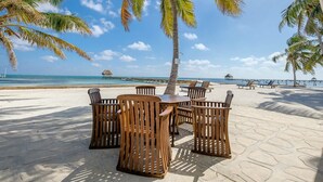 A beachside dining area with wooden tables and chairs, palm trees, and a view of the ocean and pier. - Sunset Beach "B1" - Beachfront At Its Best Plus Pool, Kayaks, Bikes & More 🏖 (San Pedro Town)