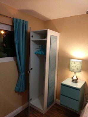 A room with a wardrobe, a bedside table with a lamp, and a window with curtains.