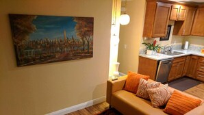 Un soggiorno con divano, un tappeto decorato, un quadro che raffigura un panorama urbano e un angolo cottura con pensili in legno e un piano di lavoro.