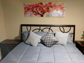 A bed with a grey quilt and two pillows, a wall with a painting of a tree, and a small table with a vase.