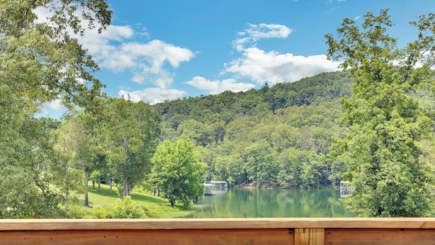 A hot tub with a wooden deck in front, overlooking a lake surrounded by lush greenery and trees.