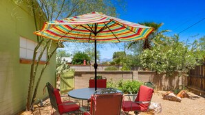 A patio with a round table and chairs, a colorful striped umbrella, and a view of a green building and trees. - Tucson Desert Rendezvous (Tucson)