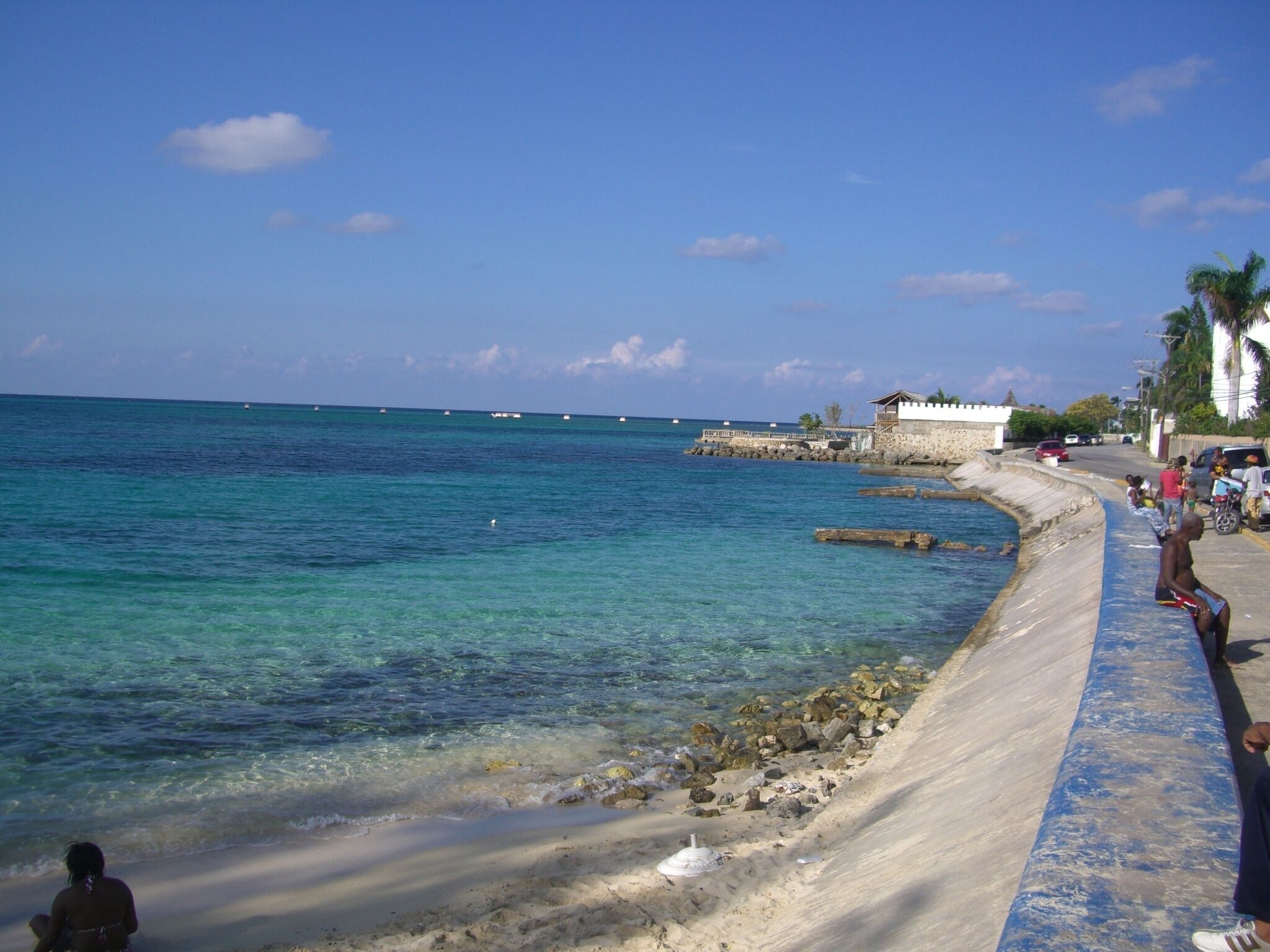 A beach with clear turquoise water, a sandy shore, and a stone wall running parallel to the water.