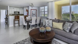 A modern living room with a gray sofa, a wooden coffee table, and a dining area with white chairs and a chandelier. - 3bed 2bath Luxury Yatch Club Condo at Aventura Fl, Parking Include! (Miami)