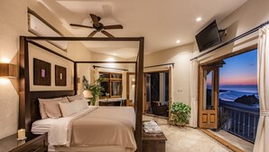 Een slaapkamer met een hemelbed, een plafondventilator, een aan de muur bevestigde televisie en een balkon met uitzicht op zee.