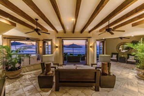 A spacious room with a wooden ceiling, a large bed, and a view of the ocean at sunset.