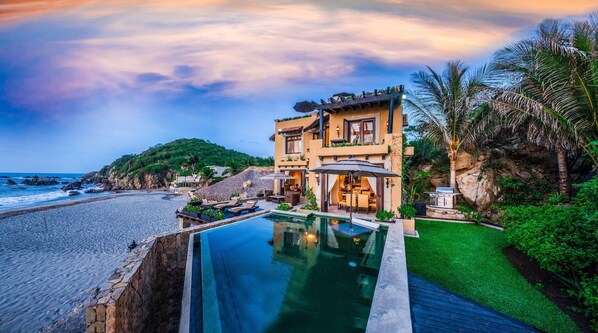 A two-story house with a swimming pool, located near a beach with a rocky cliff and palm trees.