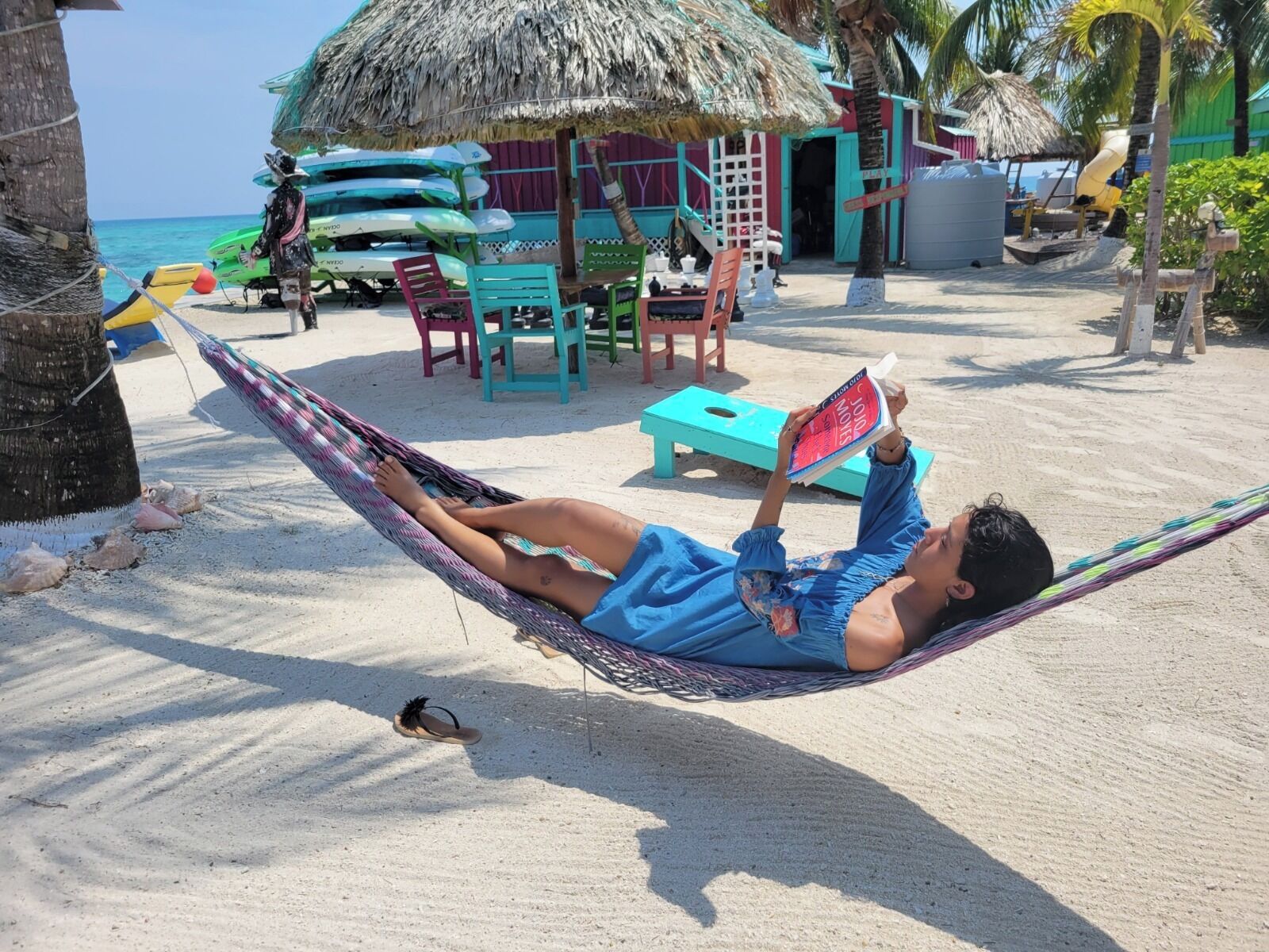 A person lying in a hammock on a sandy beach with a thatched-roof structure in the background.