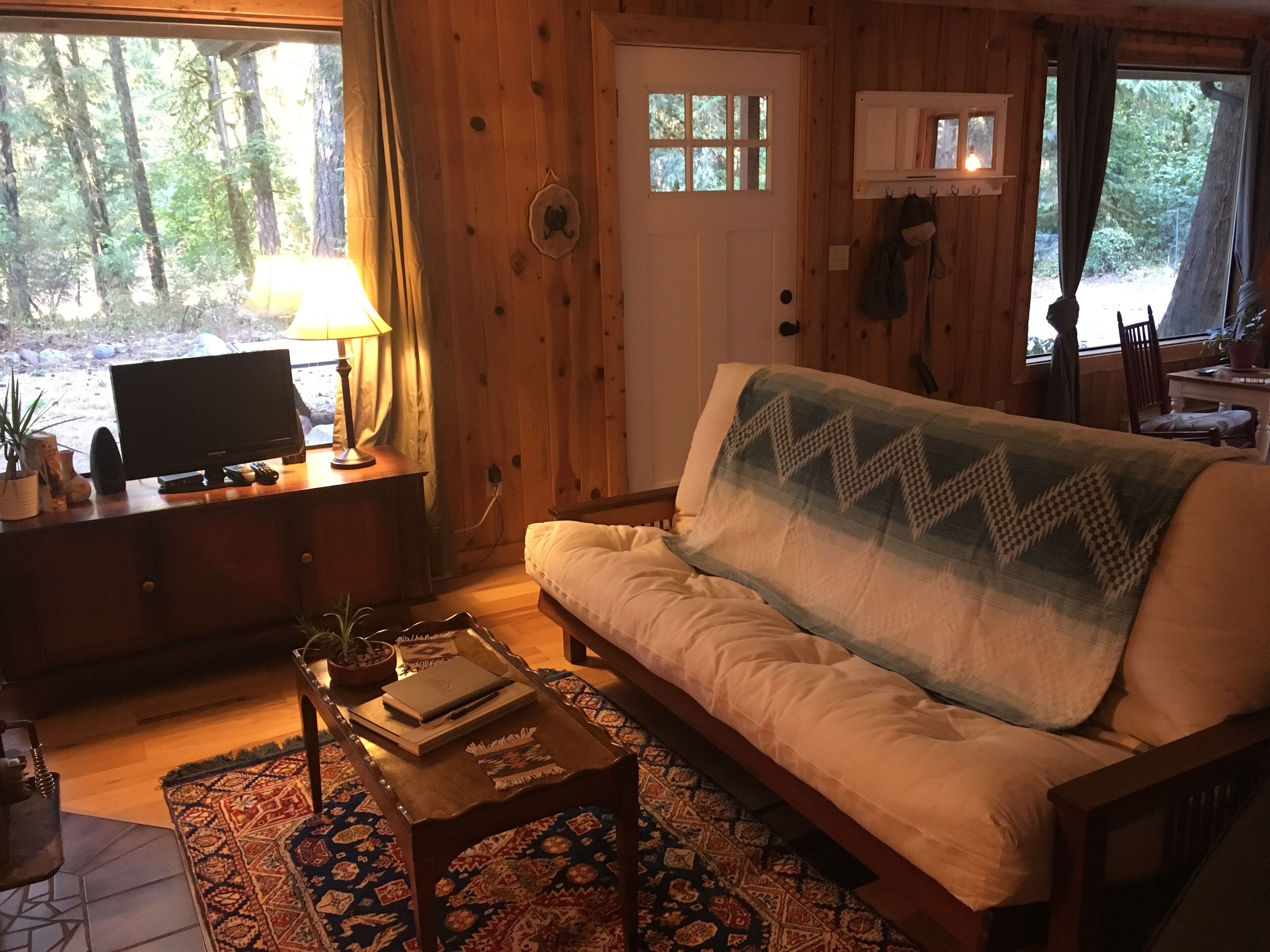 A cozy cabin interior with a sofa, a television, a lamp, and a window overlooking a forest.
