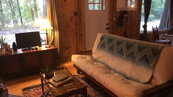 A cozy cabin interior with a sofa, a television, a lamp, and a window overlooking a forest.