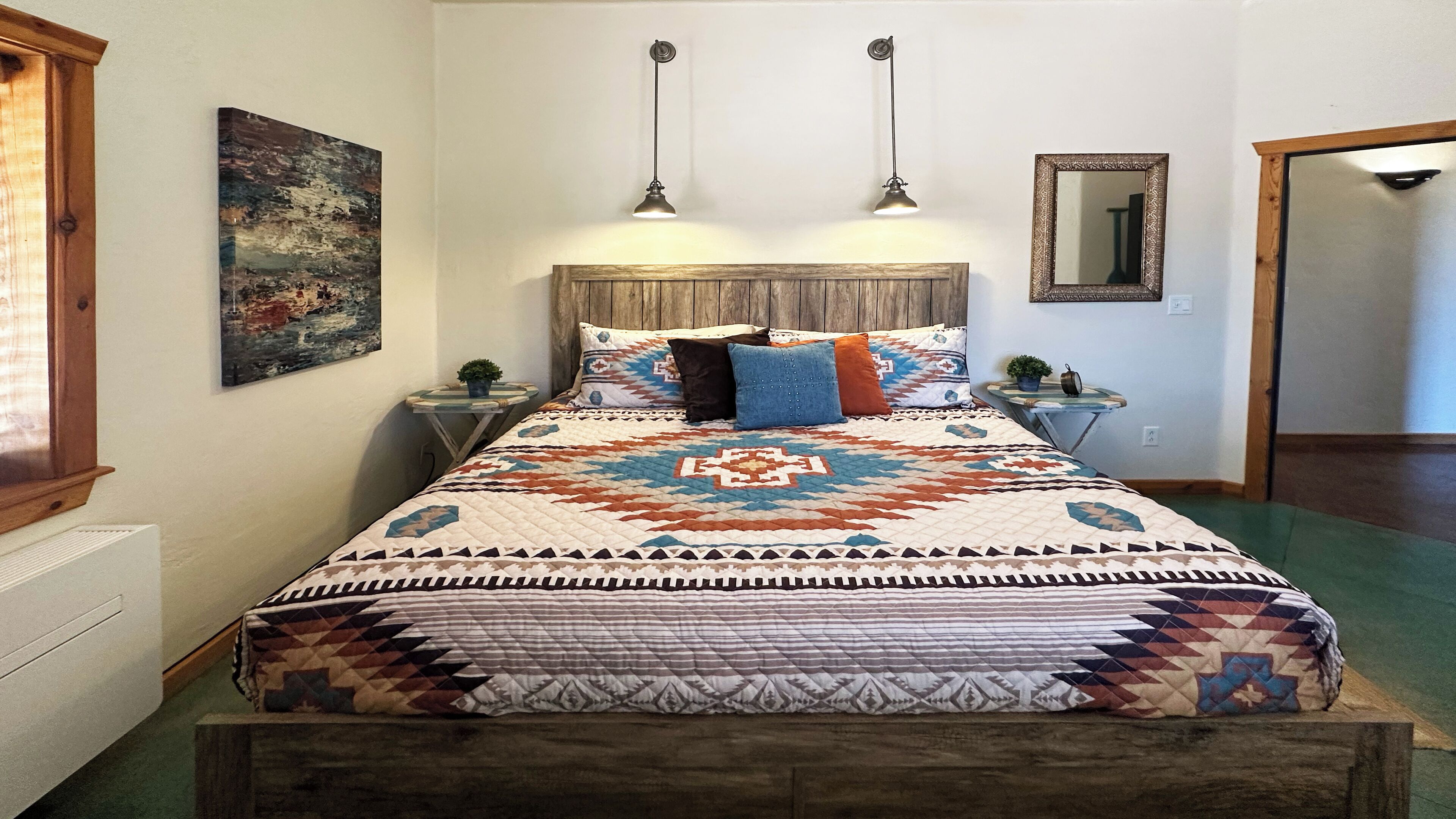 A bed with a patterned quilt, two bedside tables with lamps, a wall-mounted mirror, and a painting on the wall.