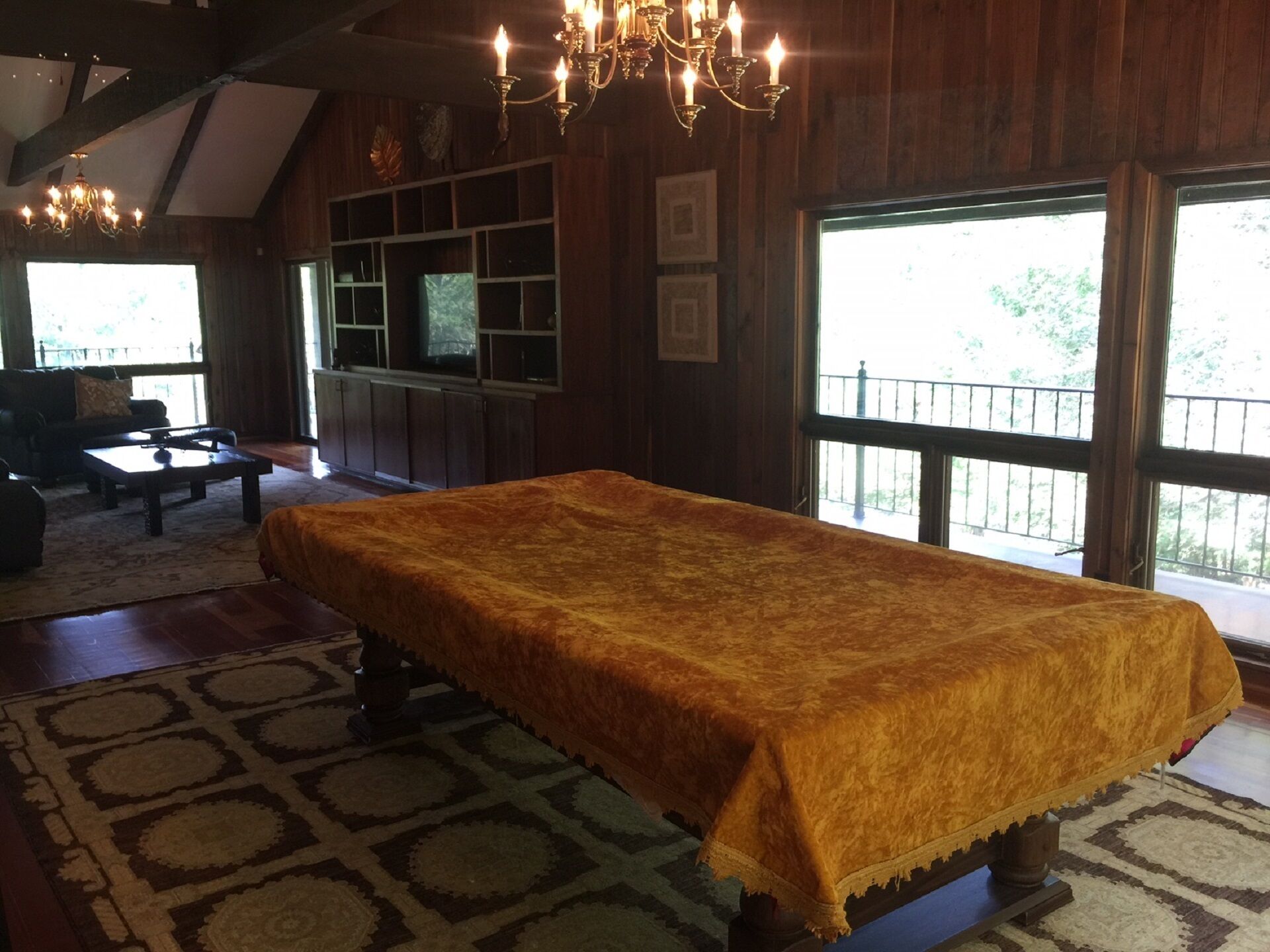 Una habitación con una mesa larga cubierta con un mantel estampado, paredes de madera, una araña, un televisor en un estante y ventanas grandes con vistas a zonas verdes.
