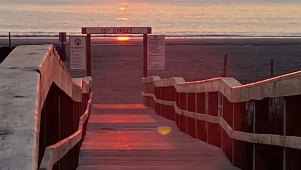 Ein hölzerner Steg führt zu einem Strand, im Hintergrund ein Sonnenuntergang.