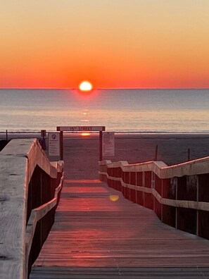 Ein hölzerner Steg führt zu einem Strand, im Hintergrund ein Sonnenuntergang.