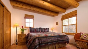 A bedroom with a wooden ceiling, a bed with a colorful blanket, two bedside lamps, a window, and a wooden chair.