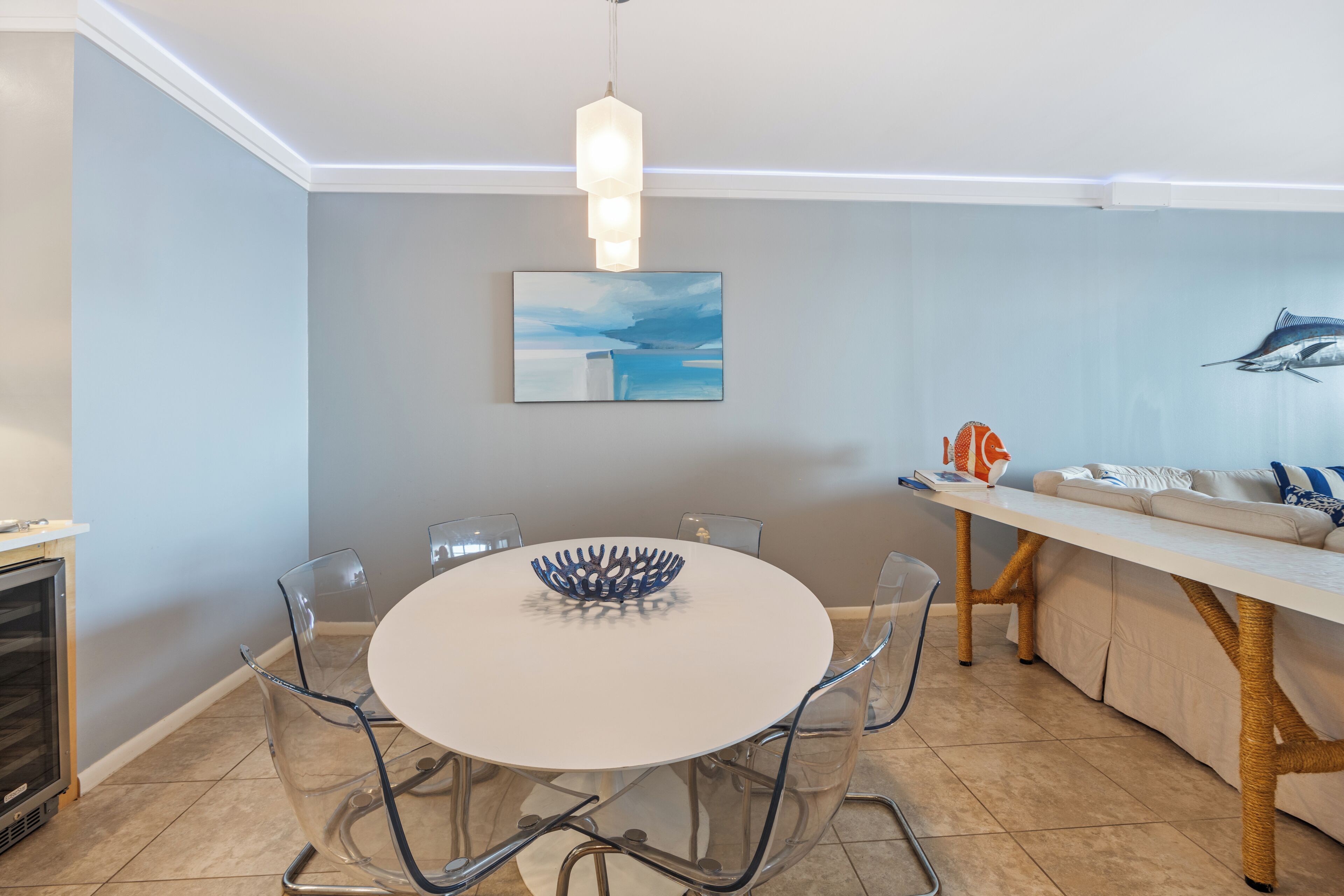 A modern dining area with a round table, clear chairs, a decorative bowl, a wall-mounted painting, and a sofa with a woven table leg.