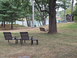 A park with three dark-colored chairs and a swing under a large tree, with a lake and a wooden structure in the background.