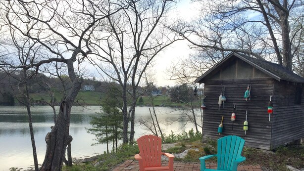 A wooden cabin with fishing equipment on the wall, two colorful Adirondack chairs, and a brick pathway leading to a lake.