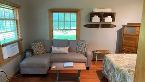 A cozy room with a grey sofa, a wooden table, a bed with floral bedding, a window with blinds, and a wooden dresser with shelves.