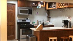 A kitchen with wooden cabinets, a stainless steel refrigerator, a microwave, a stove, and a coffee maker. There are three wooden stools at the counter.