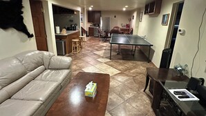 A living room with a sofa, coffee table, and ping pong table. There is a kitchen in the background with cabinets and appliances.