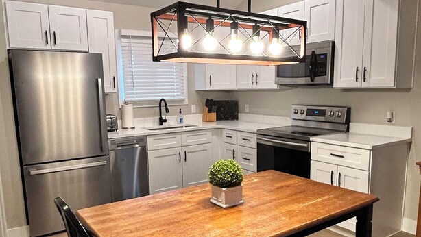 A modern kitchen with white cabinets, stainless steel appliances, a wooden dining table, and a hanging light fixture. - Fun Under The Sun, Condo 3: You Need a St. George Get-away! (St George)