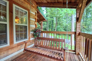A wooden porch with a swing bench, large windows, and a view of a forested area.