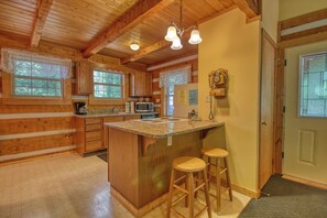 A wooden cabin interior with a kitchen featuring a central island, bar stools, and a wooden ceiling.