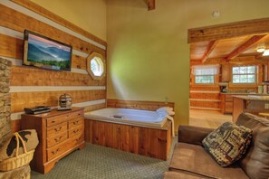 A cozy cabin interior with a wooden bathtub, a television mounted on a wall, and a kitchen area visible through an open door.