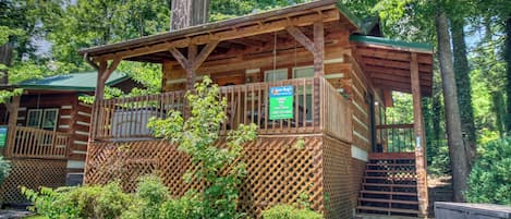 A rustic wooden cabin with a covered porch and a chimney, surrounded by lush greenery.