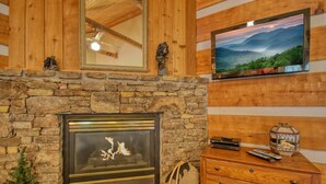 A stone fireplace with a TV mounted above it, wooden walls, and a wooden dresser with a basket and remote controls.