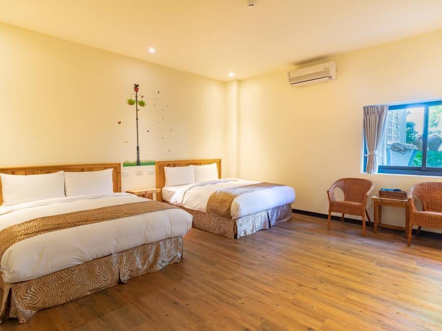 A hotel room with two beds, a wooden floor, a window with a view, wicker chairs, and an air conditioning unit.