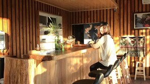 A wooden bar area with a person seated at the bar, wooden stools, and a window with blinds. - Lane 523 (Puli)