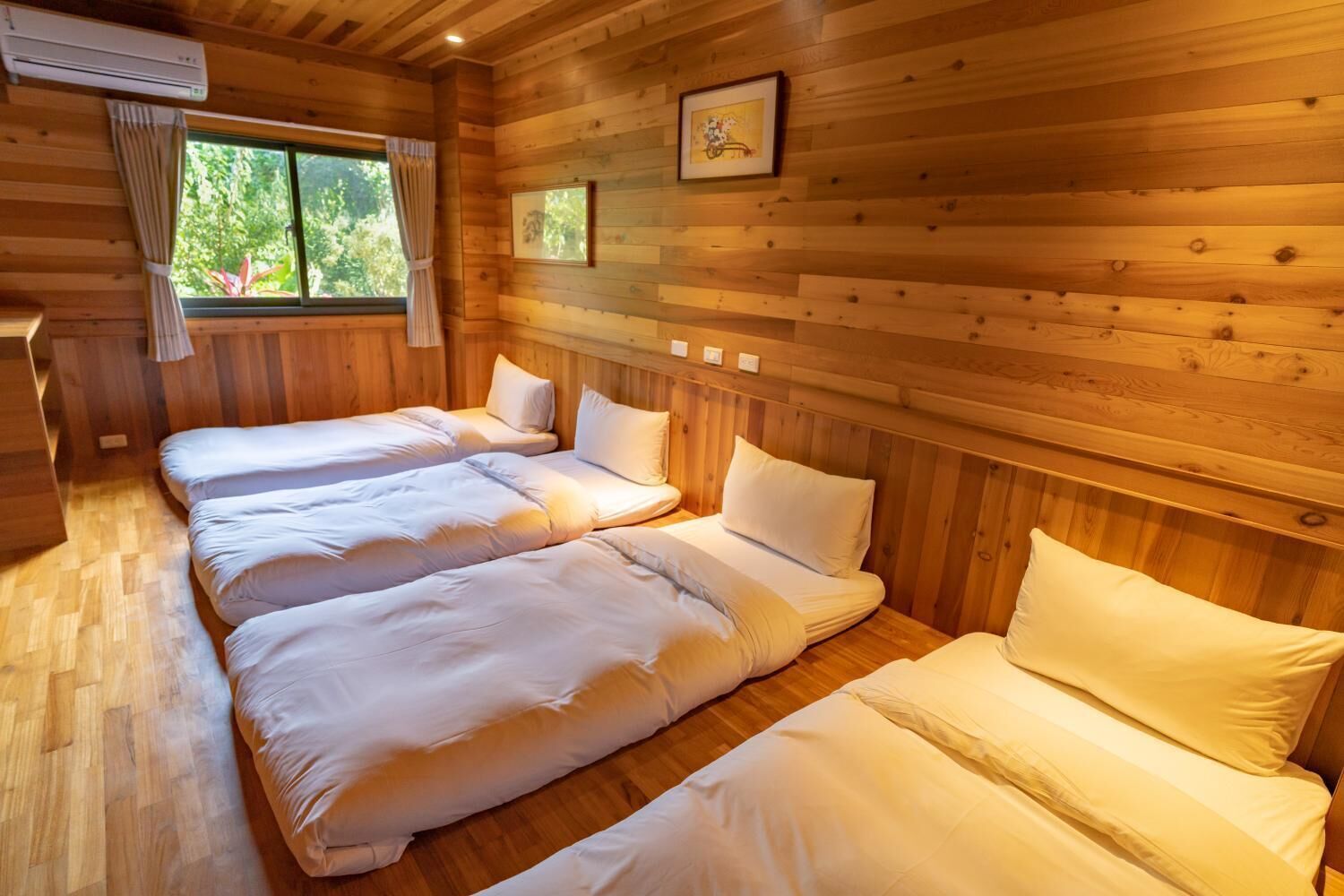A wooden cabin room with three beds, a window with curtains, and framed pictures on the wall.